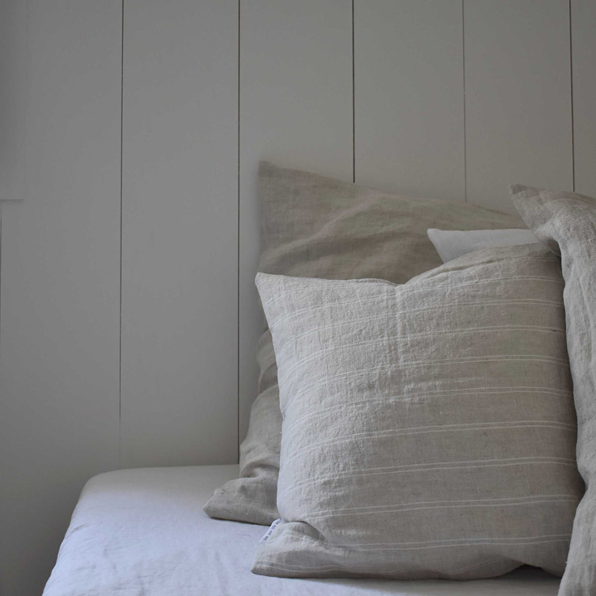 Landelijk opgemaakt bed met linnen kussenslopen in de kleur flax en pinstripe flax, tegen een witte houten panelenwand.