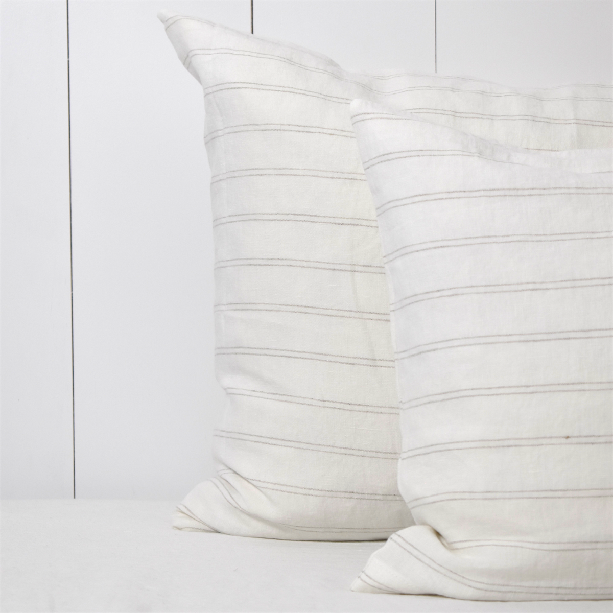 Melkwit linnen kussensloop met een dun, vlaskleurig streepje op een bed tegen een witte houten wand.