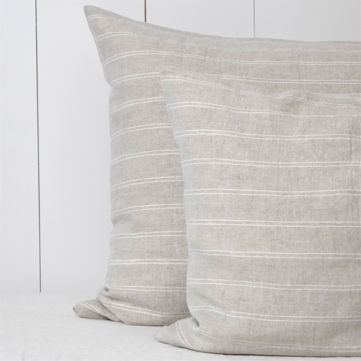 Twee linnen kussenslopen met een streepmotief op een licht bed tegen een witte houten wand.