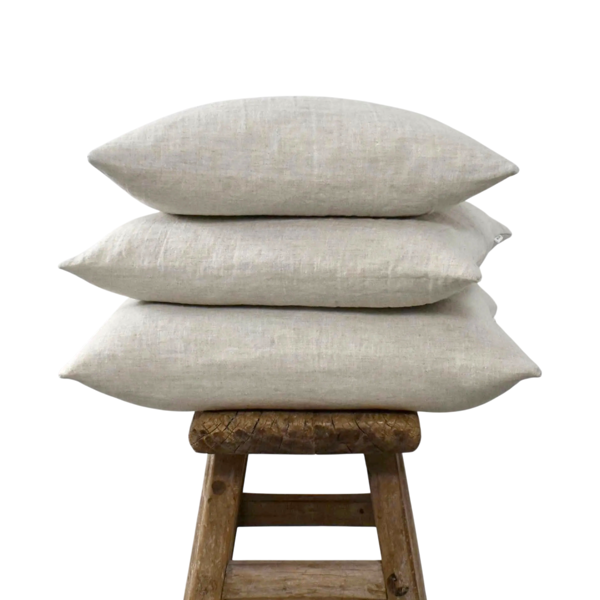 Stapel van drie licht beige linnen kussenslopen in de kleur Natural op een oud houten krukje tegen een witte achtergrond.
