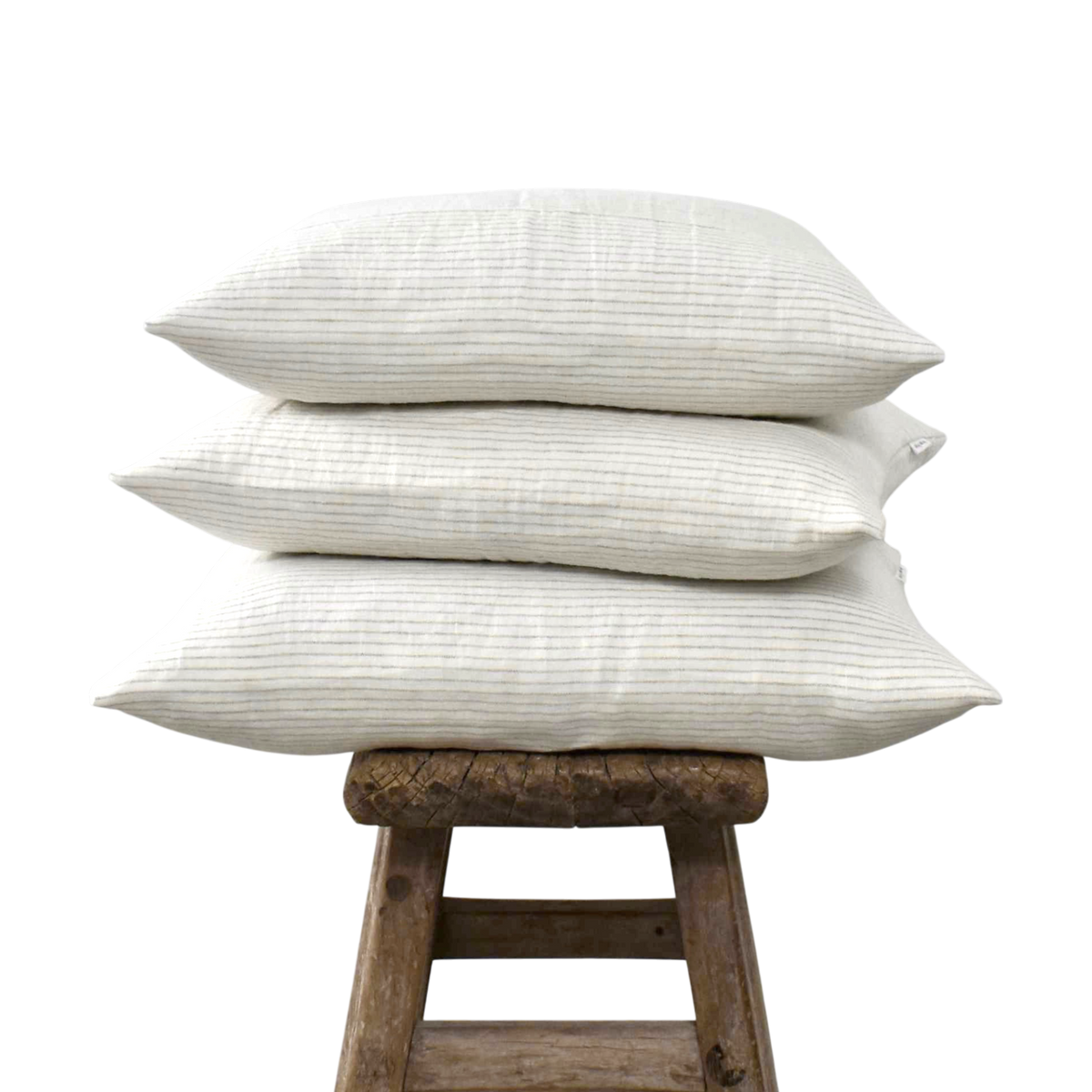 Stapel van drie linnen kussenslopen met een fijn streepmotief op een verweerd houten krukje tegen een witte achtergrond.