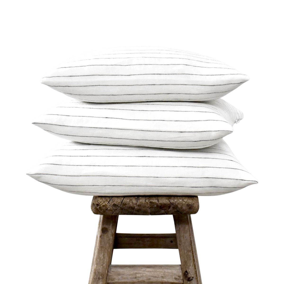 Stapel van drie witte linnen kussenslopen met een fijn donkerbruin streepje op een rustiek houten krukje tegen een witte achtergrond.