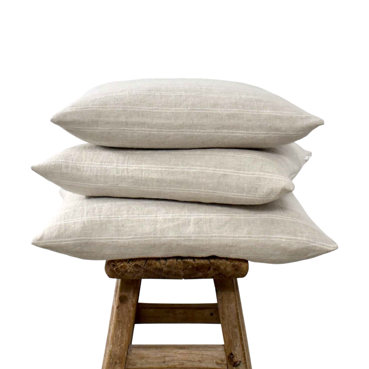 Stapel van drie beige linnen sierkussens met fijne witte strepen op een oude houten kruk.