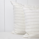 Melkwit linnen kussensloop met een dun, vlaskleurig streepje op een bed tegen een witte houten wand.