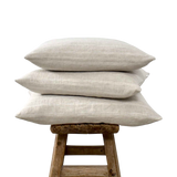 Stapel van drie beige linnen sierkussens met fijne witte strepen op een oude houten kruk.